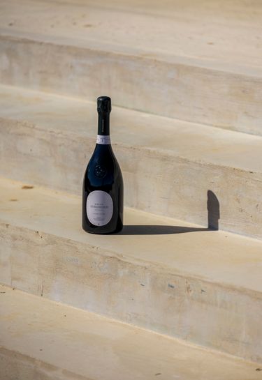 A single bottle of sparkling wine is placed on smooth beige stone steps. The bottle is dark with a prominent label reading Domaine Evremond, suggesting premium craftsmanship in English quality sparkling wine. The minimalist setting accentuates the elegance and refined presentation of the bottle.