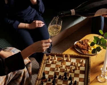 A close-up image of a person pouring sparkling wine into a glass held by another individual near a chessboard. The setting includes a wooden table with snacks, a lit candle, and a vase with greenery, suggesting a refined and cozy indoor environment. The scene conveys a quiet moment of leisure and sophistication, though no direct connection to Domaine Evremond or vineyard activity is visually evident.