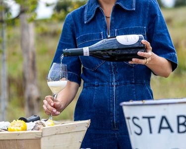 A woman in a blue denim dress pours sparkling wine into a glass outdoors, suggesting a refined tasting experience. The setting includes a rustic wooden barrel and fresh oysters, hinting at a premium English sparkling wine pairing. The scene captures an elegant and casual moment in a vineyard-like environment.