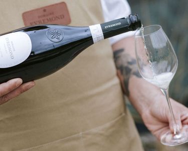 A close-up view of a person pouring Domaine Evremond English quality sparkling wine into a branded wine glass. The image highlights the careful craftsmanship and elegance associated with Domaine Evremond wines. The setting suggests a refined tasting experience celebrating premium sparkling wine.