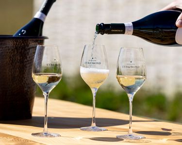 A bottle of Domaine Evremond English quality sparkling wine is being poured into a branded wine glass on a wooden table, alongside two other glasses partially filled. The scene captures premium craftsmanship associated with English sparkling wine served outdoors under natural light. The setting highlights the refined experience of Domaine Evremond wines.