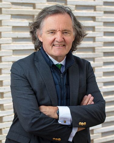 Portrait of a mature man with gray hair wearing a dark suit jacket over a black vest and white shirt standing against a textured wall background. The man is posed with arms crossed and smiling gently, presenting a confident and approachable demeanor. The image has a professional and refined atmosphere.