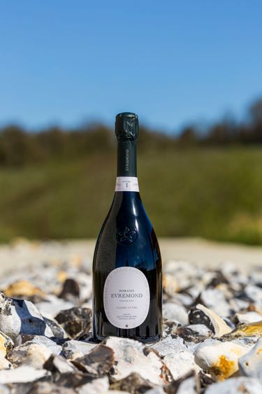 A bottle of Domaine Evremond Classic Cuvée English quality sparkling wine is displayed outdoors on a bed of stones. The image emphasizes the bottle's elegant design and label against a soft-focus natural background, highlighting premium craftsmanship. The setting sun casts natural light, enhancing the bottle's sleek appearance and the wine's refined character.