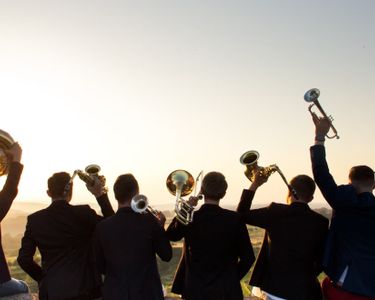 A group of musicians form a brass band playing trumpets, saxophones, and other brass instruments outdoors during sunset. The scene captures the atmospheric golden light, emphasizing the silhouettes of the players against a wide open landscape. The setting suggests a celebratory or casual music performance in an open-air environment.