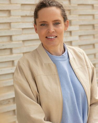 A portrait of a woman wearing a beige jacket over a light blue top, standing against a light brick wall background. The image captures a natural and relaxed expression with soft lighting highlighting her features. The neutral tones of the jacket contrast subtly with the pastel blue shirt, creating a calm and approachable atmosphere.