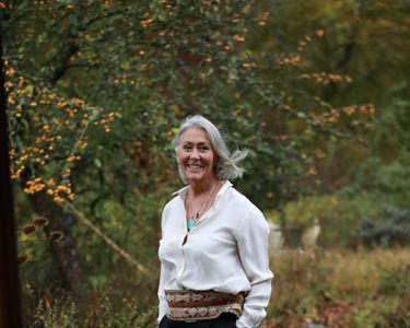 Marian Boswall stands on a stone pathway surrounded by lush greenery and autumnal foliage. She wears a white blouse paired with black wide-leg trousers and a decorative belt. The natural setting features trees with orange berries and a peaceful garden ambiance.