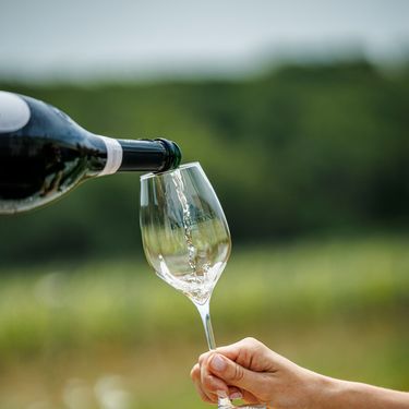 A hand holding a clear wine glass is being filled with sparkling wine from a bottle. The glass bears the Domaine Evremond logo, indicating premium English sparkling wine. The blurred background suggests a vineyard setting, highlighting a refined wine tasting experience.
