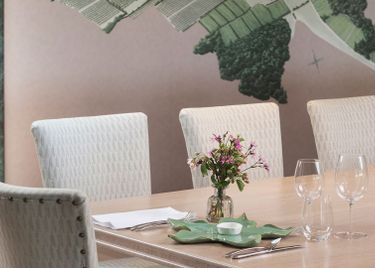 A refined meeting room featuring a long wooden table with four upholstered white chairs. The table is decorated with a small vase of fresh flowers, glassware, and neatly arranged cutlery. A large wall map providing a subtle backdrop enhances the sophisticated, professional atmosphere.