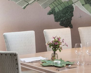 A refined meeting room featuring a long wooden table with four upholstered white chairs. The table is decorated with a small vase of fresh flowers, glassware, and neatly arranged cutlery. A large wall map providing a subtle backdrop enhances the sophisticated, professional atmosphere.