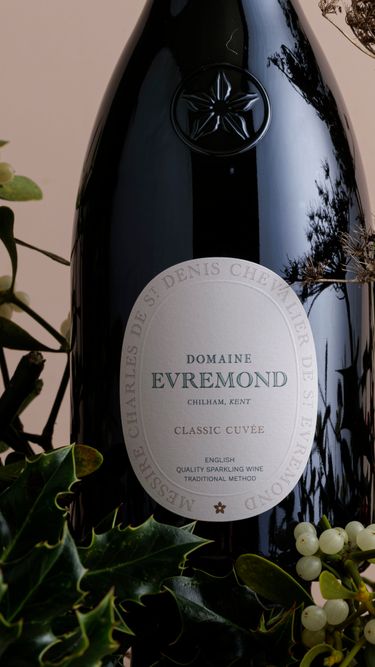 A close-up view of a bottle of Domaine Evremond Classic Cuvée English quality sparkling wine, surrounded by festive greenery and berries. The label highlights the premium craftsmanship of this traditional method sparkling wine from Chilham, Kent. The natural foliage adds a refined, seasonal atmosphere to the elegant presentation.