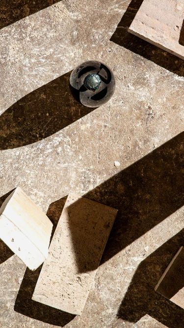 A composition of scattered light-colored bricks and a dark spherical object cast strong shadows on a textured surface. The image emphasizes the contrast between light and shadow, with an abstract, geometric arrangement. No direct connection to wine or vineyard settings is visible.