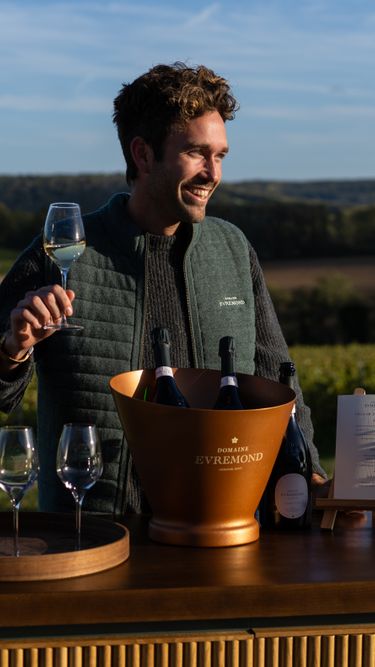 A man enjoys a glass of Domaine Evremond English quality sparkling wine during a tasting event set against a lush vineyard landscape. The scene highlights premium craftsmanship with bottles displayed in an elegant ice bucket and multiple glasses ready for tasting on a wooden table. The natural light and expansive vineyard backdrop emphasize the connection to an English vineyard setting.