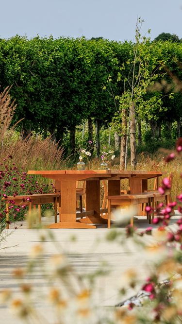 A wooden outdoor dining table with benches is set along a paved pathway surrounded by lush green trees and blooming flowers. The scene suggests a peaceful garden setting ideal for relaxation or gatherings, with natural elements enhancing the ambiance. This setting could be part of an estate's landscaped grounds, merging refined outdoor living with natural beauty.