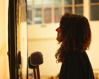 A woman is captured in profile as she strikes a large gong with a mallet in a softly lit indoor setting. Her curly hair and dark clothing contrast with the warm background light, creating an intimate atmosphere. The image focuses on the moment of musical engagement, highlighting the reflective surface of the gong and the contemplative expression of the woman.