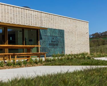 Modern exterior of Domaine Evremond tasting room featuring a textured cream brick facade and a green stone sign with the estate name. The setting includes outdoor wooden benches and natural greenery with a backdrop of rural English countryside. This refined architecture complements the premium craftsmanship associated with Domaine Evremond English quality sparkling wine.