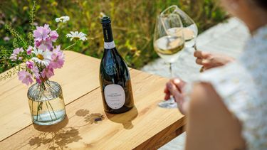 An outdoor setting featuring a bottle of Domaine Evremond English quality sparkling wine on a wooden table beside a vase of fresh flowers. Two glasses of sparkling wine are held by a person, suggesting a relaxed wine tasting experience. The scene reflects premium craftsmanship and the estate’s vineyard ambiance.