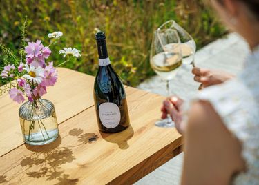 An outdoor setting featuring a bottle of Domaine Evremond English quality sparkling wine on a wooden table beside a vase of fresh flowers. Two glasses of sparkling wine are held by a person, suggesting a relaxed wine tasting experience. The scene reflects premium craftsmanship and the estate’s vineyard ambiance.