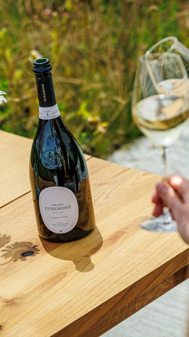 An outdoor setting featuring a bottle of Domaine Evremond English quality sparkling wine on a wooden table beside a vase of fresh flowers. Two glasses of sparkling wine are held by a person, suggesting a relaxed wine tasting experience. The scene reflects premium craftsmanship and the estate’s vineyard ambiance.