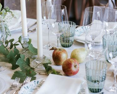 A refined dining table arrangement featuring glassware, white taper candles in crystal holders, fresh apples and a pear, and floral greenery on a white textured tablecloth. The setting conveys an atmosphere of sophistication and attention to detail, suitable for intimate gatherings or celebratory meals. The natural tones and delicate elements evoke a calm, inviting ambiance.