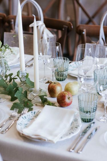 A refined dining table arrangement featuring glassware, white taper candles in crystal holders, fresh apples and a pear, and floral greenery on a white textured tablecloth. The setting conveys an atmosphere of sophistication and attention to detail, suitable for intimate gatherings or celebratory meals. The natural tones and delicate elements evoke a calm, inviting ambiance.
