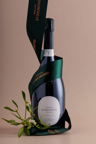 A bottle of Domaine Evremond Classic Cuvée English quality sparkling wine elegantly displayed with a dark green ribbon branded with the estate's name. The bottle features a refined label highlighting its premium craftsmanship and traditional method. A sprig of mistletoe nearby adds a seasonal, sophisticated touch against a soft beige background.