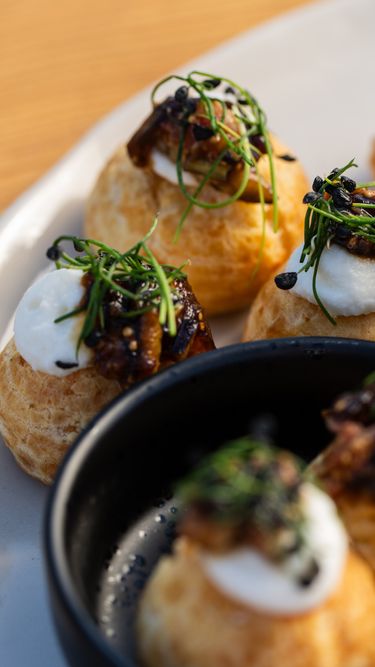 Close-up of gourmet savory canapés featuring puff pastry bases topped with creamy white cheese, sautéed mushrooms, herbs, and seasoning. The detailed focus highlights the texture and freshness of the ingredients, presented on a light-colored plate and a black bowl. This refined appetizer showcases intricate culinary craftsmanship suitable for elegant dining occasions.