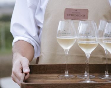 A person wearing a beige apron branded with Domaine Evremond is holding a wooden tray with four glasses of English quality sparkling wine. The setting suggests an elegant tasting experience highlighting premium craftsmanship associated with Domaine Evremond. The background indicates an outdoor vineyard setting, reinforcing the estate's connection to fine English sparkling wine production.