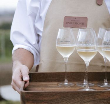 A person wearing a beige apron branded with Domaine Evremond is holding a wooden tray with four glasses of English quality sparkling wine. The setting suggests an elegant tasting experience highlighting premium craftsmanship associated with Domaine Evremond. The background indicates an outdoor vineyard setting, reinforcing the estate's connection to fine English sparkling wine production.
