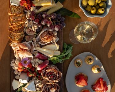 An elegant charcuterie board featuring assorted cheeses, cured meats, grapes, nuts, cherry tomatoes, and crackers, accompanied by a plate of smoked salmon canapés and a bowl of green olives. Two glasses of white sparkling wine and a wine bottle are visible, suggesting a refined tasting experience with Domaine Evremond English quality sparkling wine. The setting is bathed in warm natural light on a wooden table, highlighting premium craftsmanship and a sophisticated presentation.
