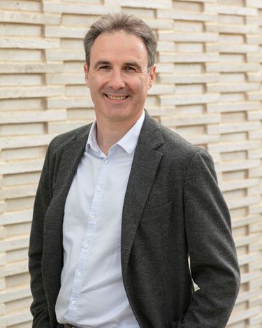 A smiling man dressed in a gray blazer and white shirt stands against a textured beige brick wall. The portrait captures a neutral professional and approachable demeanor in a well-lit outdoor setting.