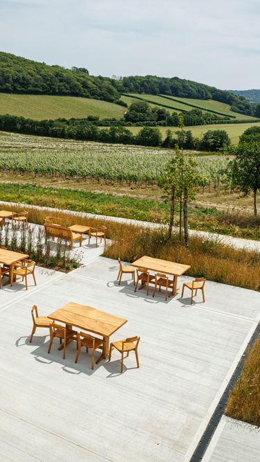 A peaceful outdoor seating area featuring wooden tables and chairs set on a concrete patio overlooks a rural landscape with rolling fields and dense tree lines. The natural surroundings include a mix of grassy areas and cultivated land. The scene conveys a serene atmosphere ideal for relaxation or casual gatherings.