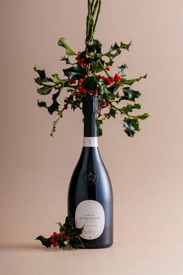 A bottle of Domaine Evremond Classic Cuvée English quality sparkling wine is elegantly presented with festive holly branches and red berries. The composition highlights premium craftsmanship and seasonal refinement, set against a neutral beige background. This image reflects the sophistication associated with Domaine Evremond’s sparkling wines.