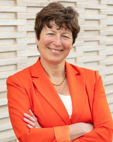 Portrait of a middle-aged woman wearing an orange blazer and a gold necklace, standing against a textured beige brick wall. The image emphasizes professional confidence and approachability with a neutral background. Suitable for business or personal branding contexts.