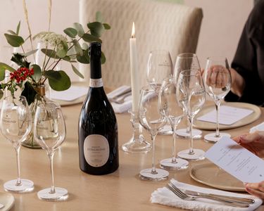 An elegant table setting featuring a bottle of Domaine Evremond English quality sparkling wine surrounded by multiple branded wine glasses. The scene includes a lit candle, floral arrangements, and people seated for a refined tasting experience. The arrangement highlights premium craftsmanship and a sophisticated ambiance ideal for wine tasting events.