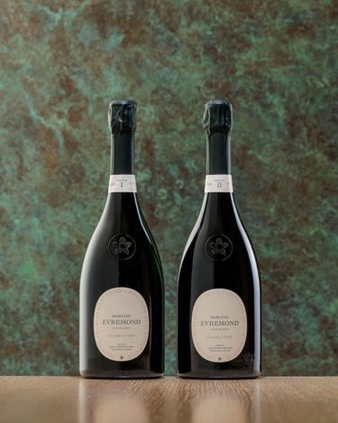 Two bottles of Domaine Evremond Classic Cuvée English quality sparkling wine are displayed against a textured green and brown backdrop on a light wood surface. The premium craftsmanship of the sparkling wine is evident in the elegant bottle design and subtle label details. This image highlights refined presentation suitable for an upscale wine setting.