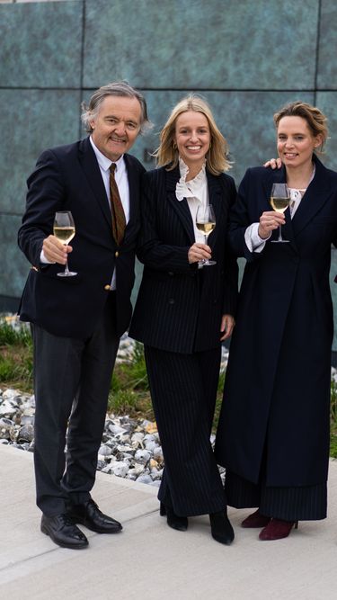 Four individuals dressed in formal attire stand outdoors on a paved area holding glasses of white wine. The group appears to be enjoying a casual celebratory moment against a modern textured wall with a small rock garden edge. The scene conveys a refined yet relaxed atmosphere suitable for a professional or social gathering.