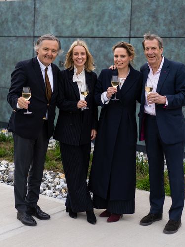 Four individuals dressed in formal attire stand outdoors on a paved area holding glasses of white wine. The group appears to be enjoying a casual celebratory moment against a modern textured wall with a small rock garden edge. The scene conveys a refined yet relaxed atmosphere suitable for a professional or social gathering.