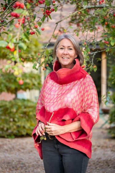 Marian Boswall stands outdoors in a garden setting, surrounded by branches bearing small red berries. She is wearing a red patterned shawl and holding some berries in her hands, with a blurred background of foliage and a building. The scene conveys a natural, serene atmosphere.