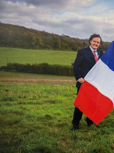 Pierre-Emmanuel Taittinger and Patrick McGrath stand in a grassy rural field holding the French and Union Jack flags. The outdoor scene suggests a countryside setting with rolling hills in the background under a cloudy sky. The image conveys a sense of cooperation or celebration between French and British interests.