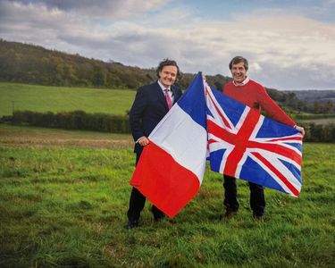 Pierre-Emmanuel Taittinger and Patrick McGrath stand in a grassy rural field holding the French and Union Jack flags. The outdoor scene suggests a countryside setting with rolling hills in the background under a cloudy sky. The image conveys a sense of cooperation or celebration between French and British interests.