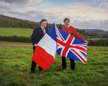 Two men stand in a grassy rural field holding the French and Union Jack flags. The outdoor scene suggests a countryside setting with rolling hills in the background under a cloudy sky. The image conveys a sense of cooperation or celebration between French and British interests.