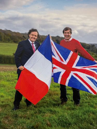 Two men stand in a grassy rural field holding the French and Union Jack flags. The outdoor scene suggests a countryside setting with rolling hills in the background under a cloudy sky. The image conveys a sense of cooperation or celebration between French and British interests.