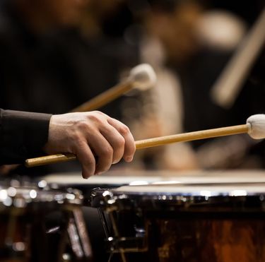 A close-up view of a musician's hand holding a mallet and playing timpani drums during a performance. The scene highlights the craftsmanship of the percussion instrument and the focus of the percussionist. The background is softly blurred, emphasizing the drum and hand in the foreground.
