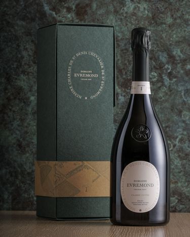 A bottle of Domaine Evremond Classic Cuvée English quality sparkling wine stands beside its elegant box, both featuring refined branding. The dark green and gold packaging complements the premium craftsmanship of this sparkling wine. The scene highlights the sophisticated presentation associated with Domaine Evremond sparkling wines.