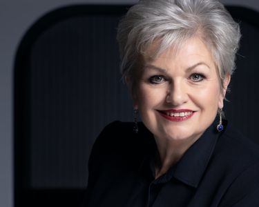 A refined portrait of a mature woman with short silver hair wearing a dark outfit and blue earrings. The image highlights her warm smile and confident expression against a dark, softly lit background. The photographic style emphasizes clarity and a professional, composed mood.