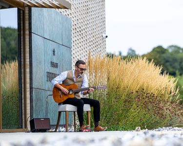 A man seated outdoors plays an acoustic guitar near a modern building with decorative paneling. The scene includes dry golden grasses and lush green foliage in the background, evoking a peaceful natural setting. The man wears casual yet smart attire including a vest and sunglasses, contributing to a relaxed, artistic atmosphere.