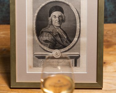 A framed black and white historical portrait of an elderly man in classical attire is displayed on a wooden surface. In front of the portrait, a glass filled with pale golden liquid is slightly out of focus. The setting suggests a refined environment blending art and a casual drink.
