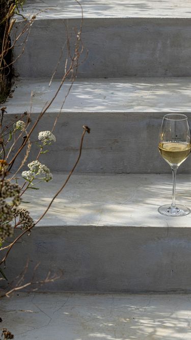 A bottle of Domaine Evremond English quality sparkling wine alongside a filled wine glass is placed on outdoor concrete steps next to wildflowers and an oyster shell. The scene suggests a refined and natural setting, highlighting premium craftsmanship and the winery's connection to its vineyard surroundings.