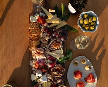 An elegant spread featuring a charcuterie board with assorted cheeses, cured meats, nuts, grapes, and crackers, complemented by a bottle and glasses of sparkling wine. Additional accompaniments include olives, bread, and small hors d'oeuvres. The natural wood table and warm lighting create a refined atmosphere suited for a sophisticated gathering.