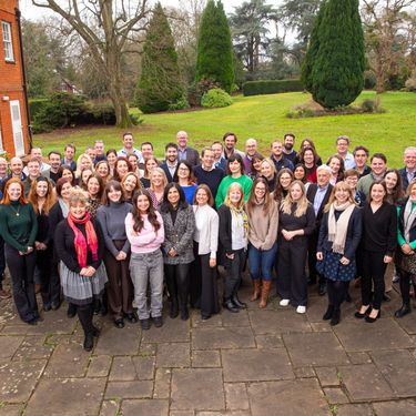 A large group of people posing together outdoors on a stone patio beside a red brick building. The setting features greenery and trees, suggesting a park or garden environment on a calm day. Everyone is dressed in smart casual attire, conveying a cheerful and social atmosphere.